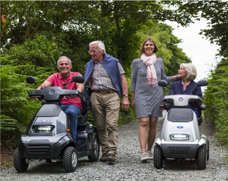 Four seniors on a scenic path: two driving mobility scooters, one wearing a pink scarf standing and smiling, and one walking with a jacket draped over his shoulders. All are enjoying a pleasant outdoor stroll surrounded by greenery and tall bushes.