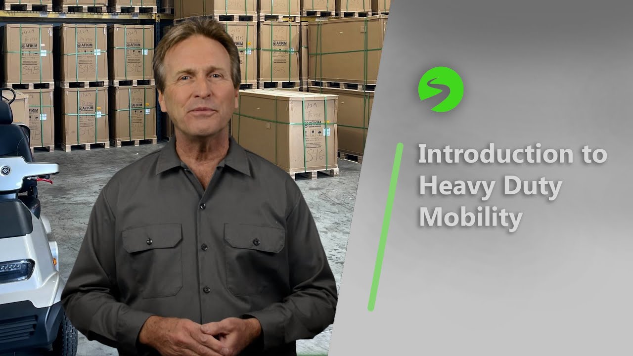 Presenter standing in warehouse with heavy-duty mobility scooters and stacked boxes, introduction to mobility solutions.